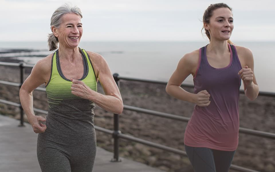 Older and younger woman running on a boardwalk by a beach
