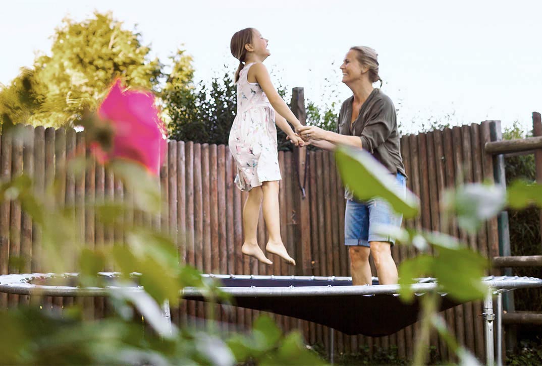 Carefree woman holds hands of laughing girl while jumping on trampoline outside.